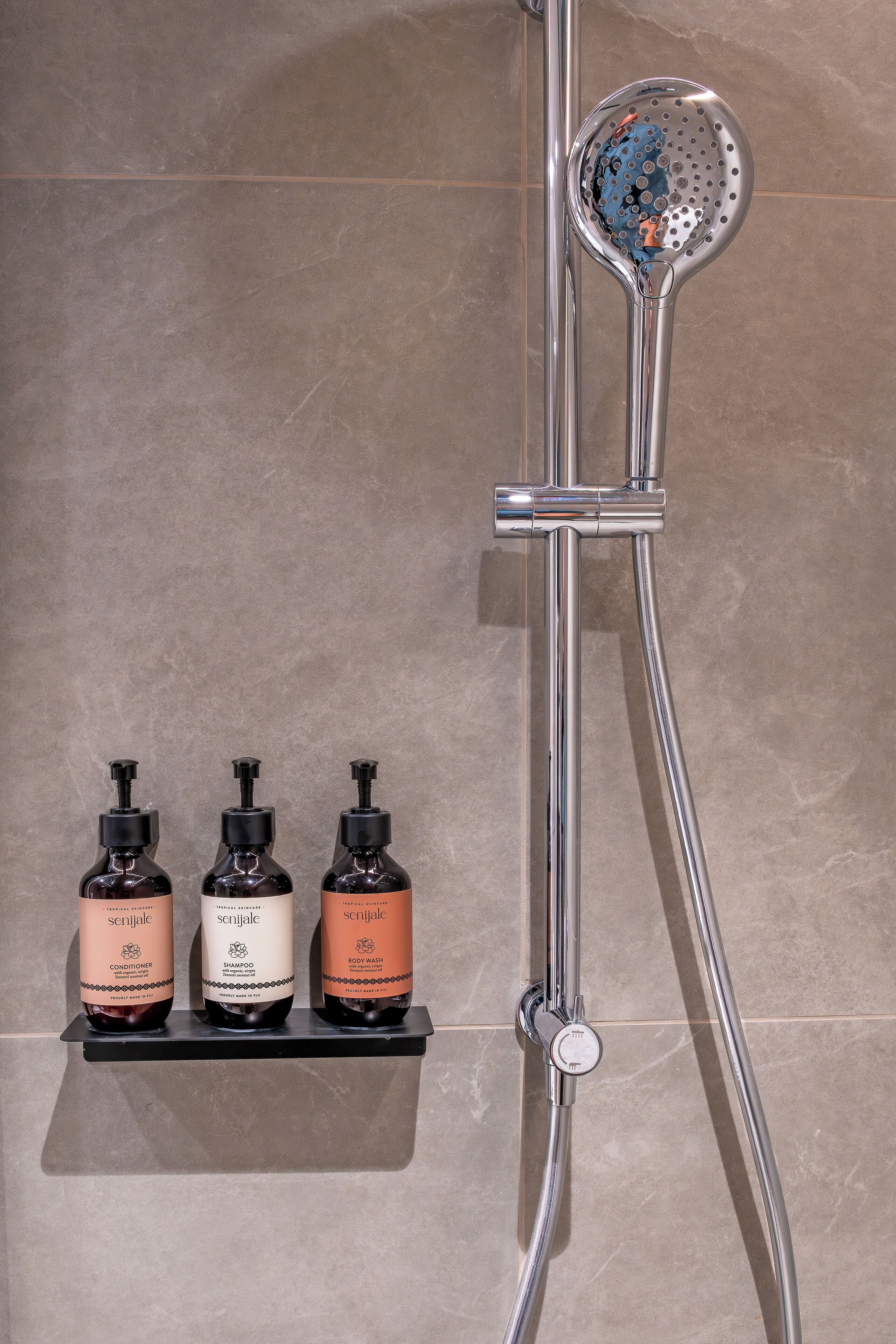 A shower with a handheld showerhead and a wall-mounted control valve. Below the shower, there are three bottles of Senjale products on a shelf.