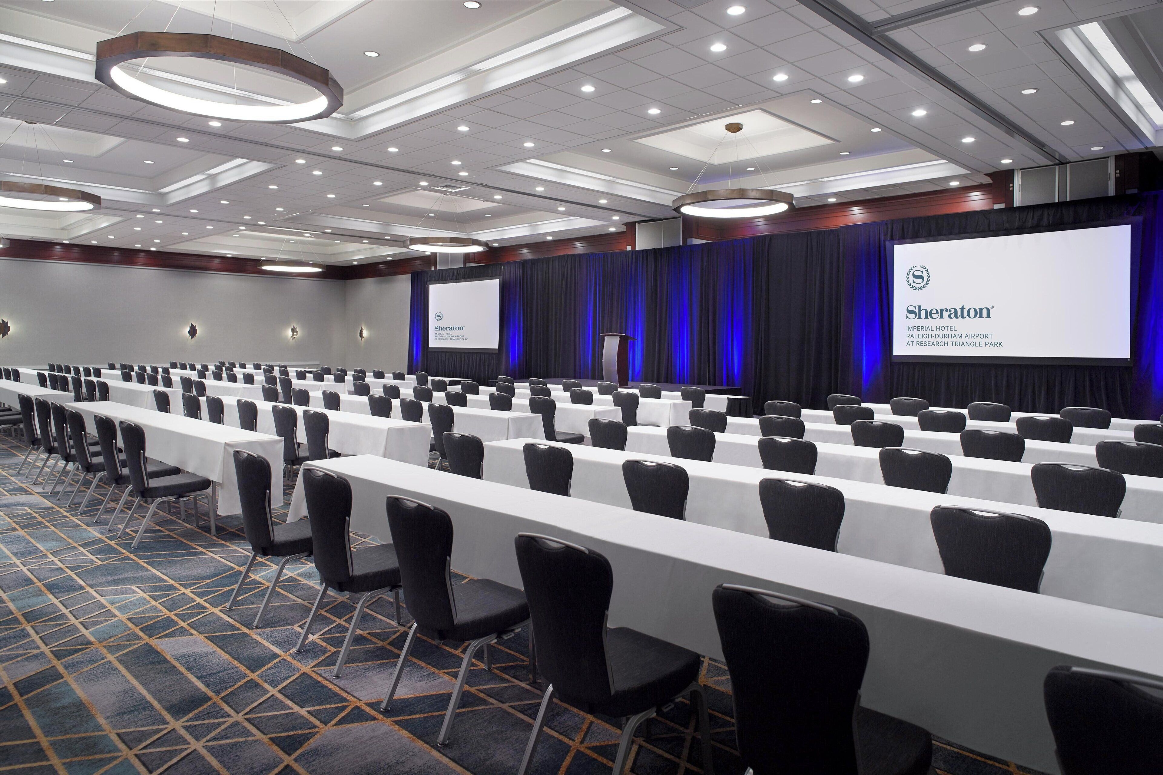 A conference room with rows of tables and chairs, a large screen displaying the Sheraton logo, and a blue curtain backdrop.