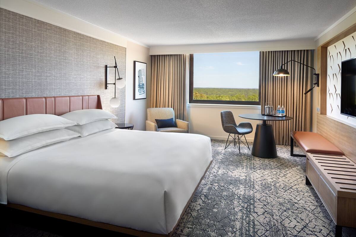 A hotel room with a large bed, a desk, a chair, a bench, a TV, and a window with a view of the landscape.