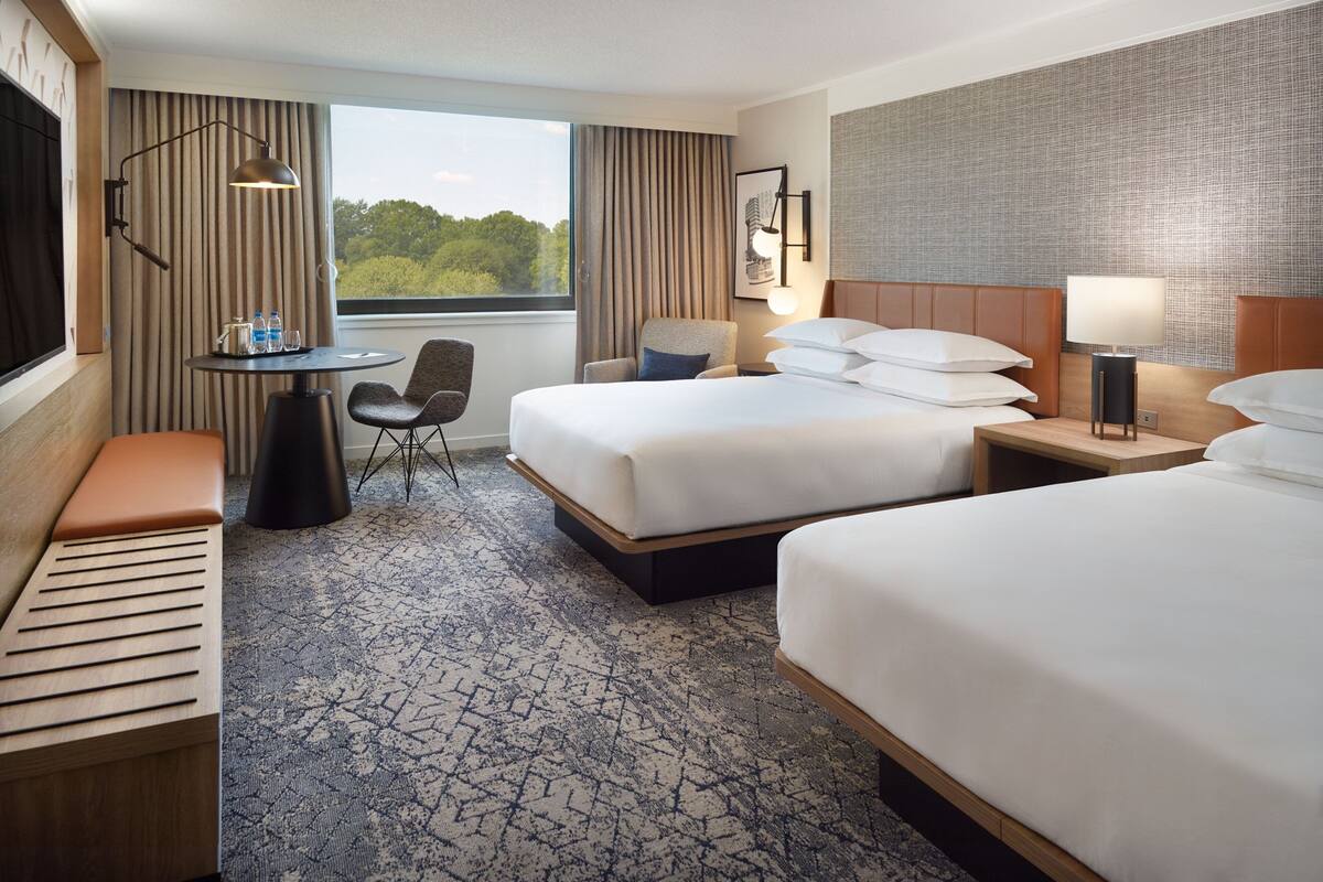 A hotel room with two beds, a desk with a chair, a small table with water bottles, and a view of greenery through the window.