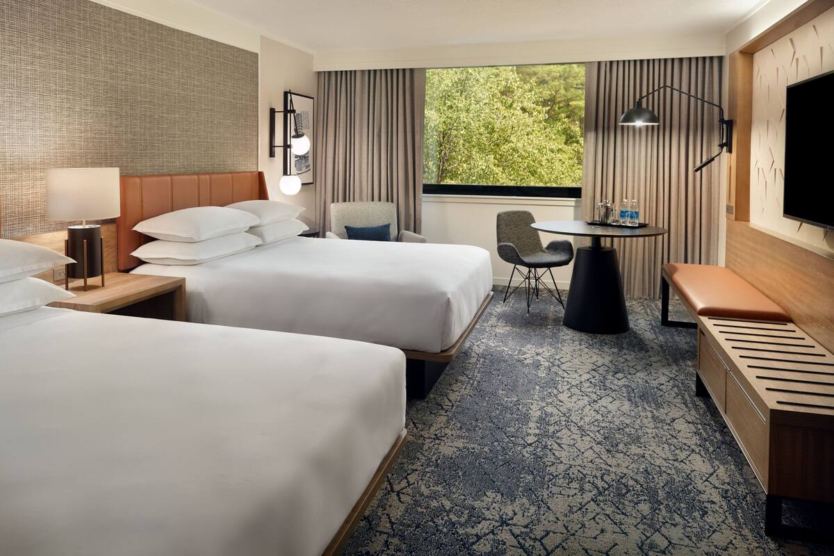 A hotel room with two beds, a desk, a chair, a TV, and a window with a view of greenery.