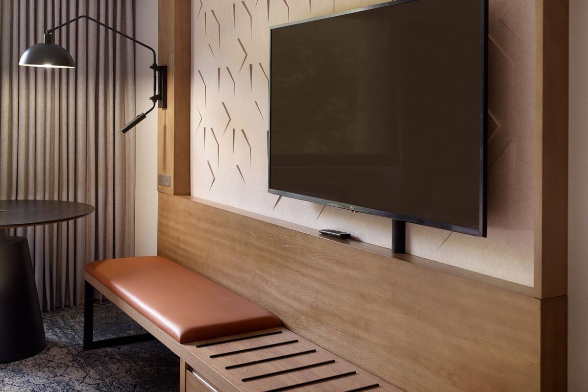 A modern hotel room with a large flat-screen TV mounted on a wooden wall, a bench, a small round table with a lamp, and a patterned wall.