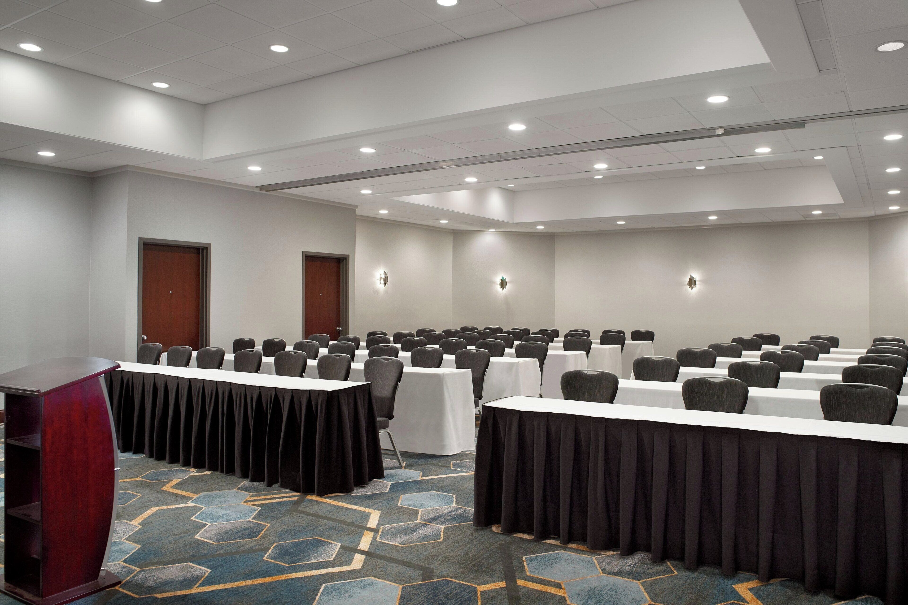 A conference room with rows of tables and chairs, a podium, and a carpeted floor.