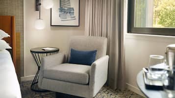 A hotel room with a patterned armchair, a small side table, a framed picture on the wall, a window with curtains, and a bed with pillows.