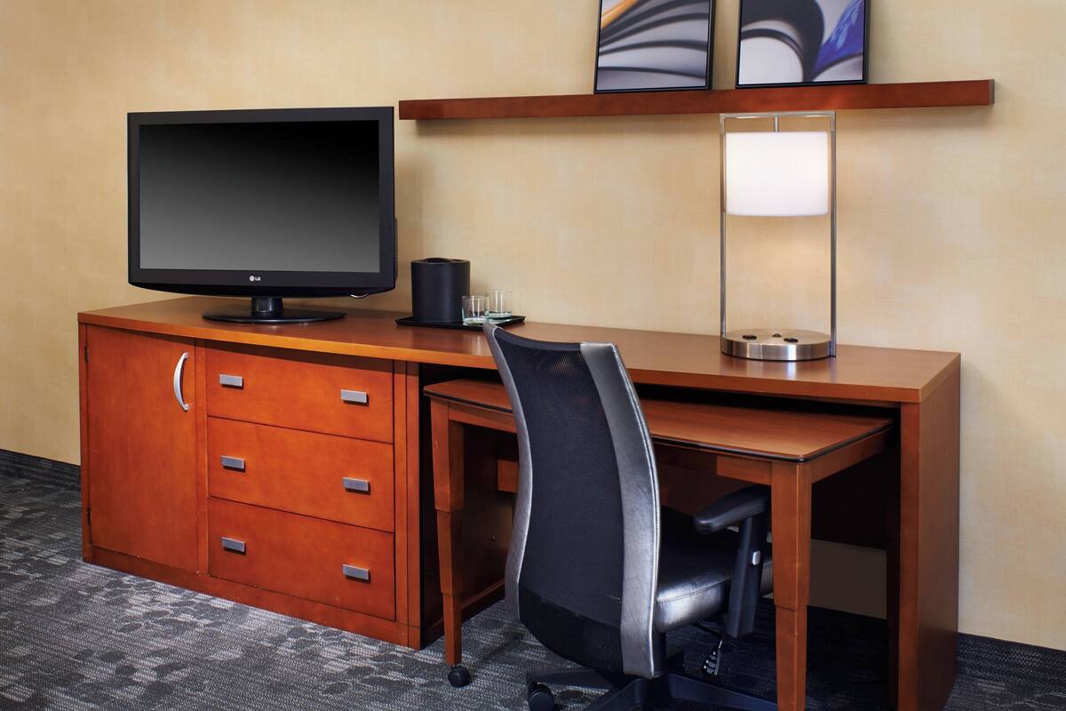 A hotel room with a wooden desk, a black office chair, a flat-screen TV, a lamp, and a minibar.