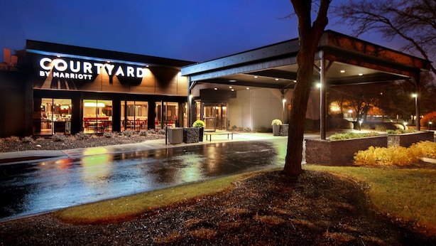 Ein Courtyard by Marriott Hotel im Abenddämmerung mit nasser Fußgängerzone, Außenbestuhlung und einem überdachten Bereich.