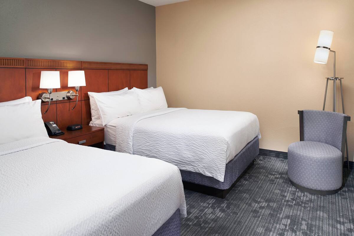 A hotel room with two beds, a nightstand with a phone, a lamp, and a round ottoman.