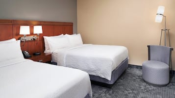 A hotel room with two beds, a nightstand with a phone, a lamp, and a round ottoman.