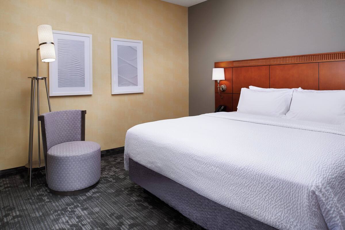 A hotel room with a large bed, a round ottoman, two framed artworks on the wall, and a floor lamp.