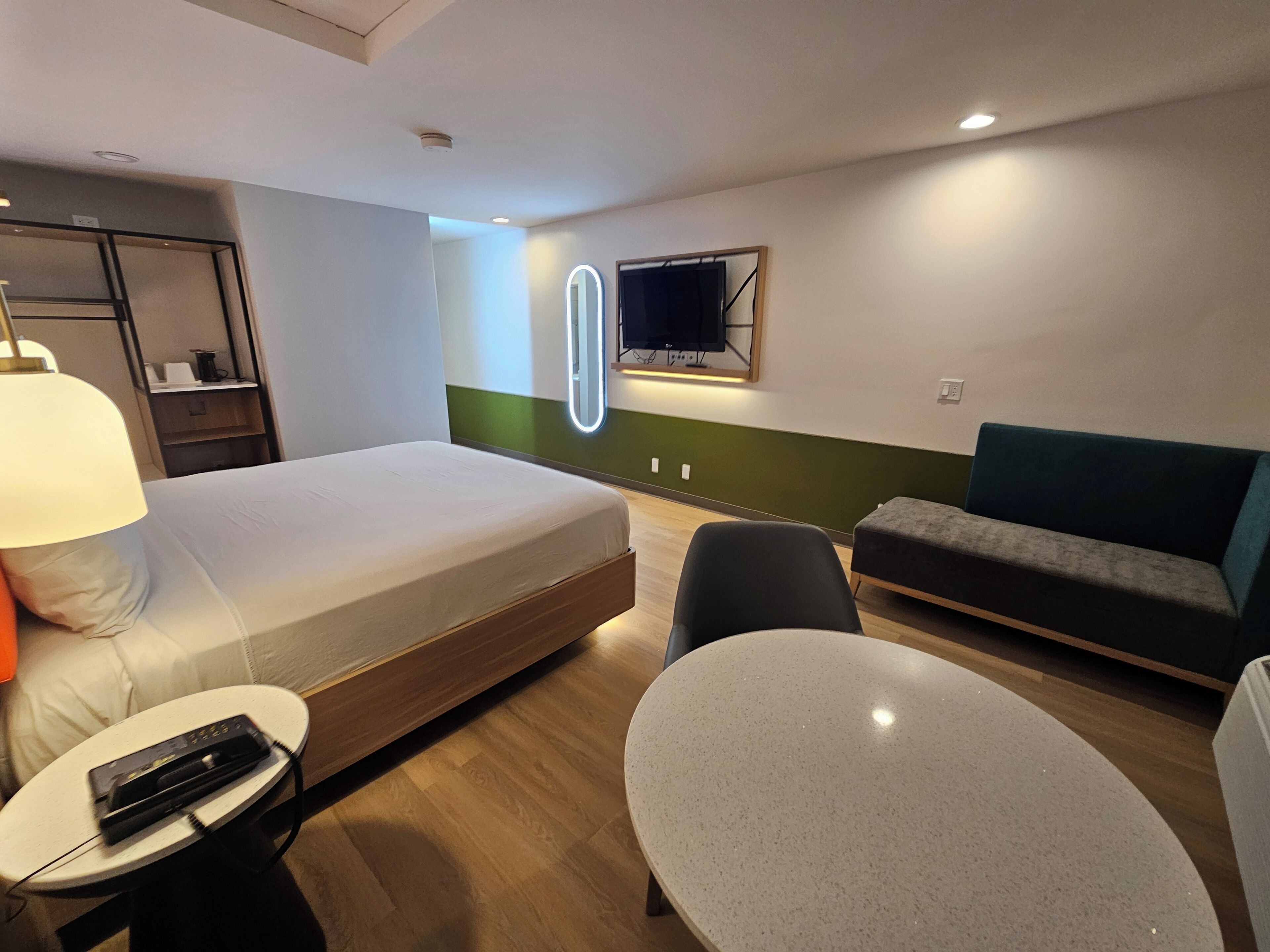 A hotel room with a bed, a desk, a chair, a sofa, a TV, and a mirror.