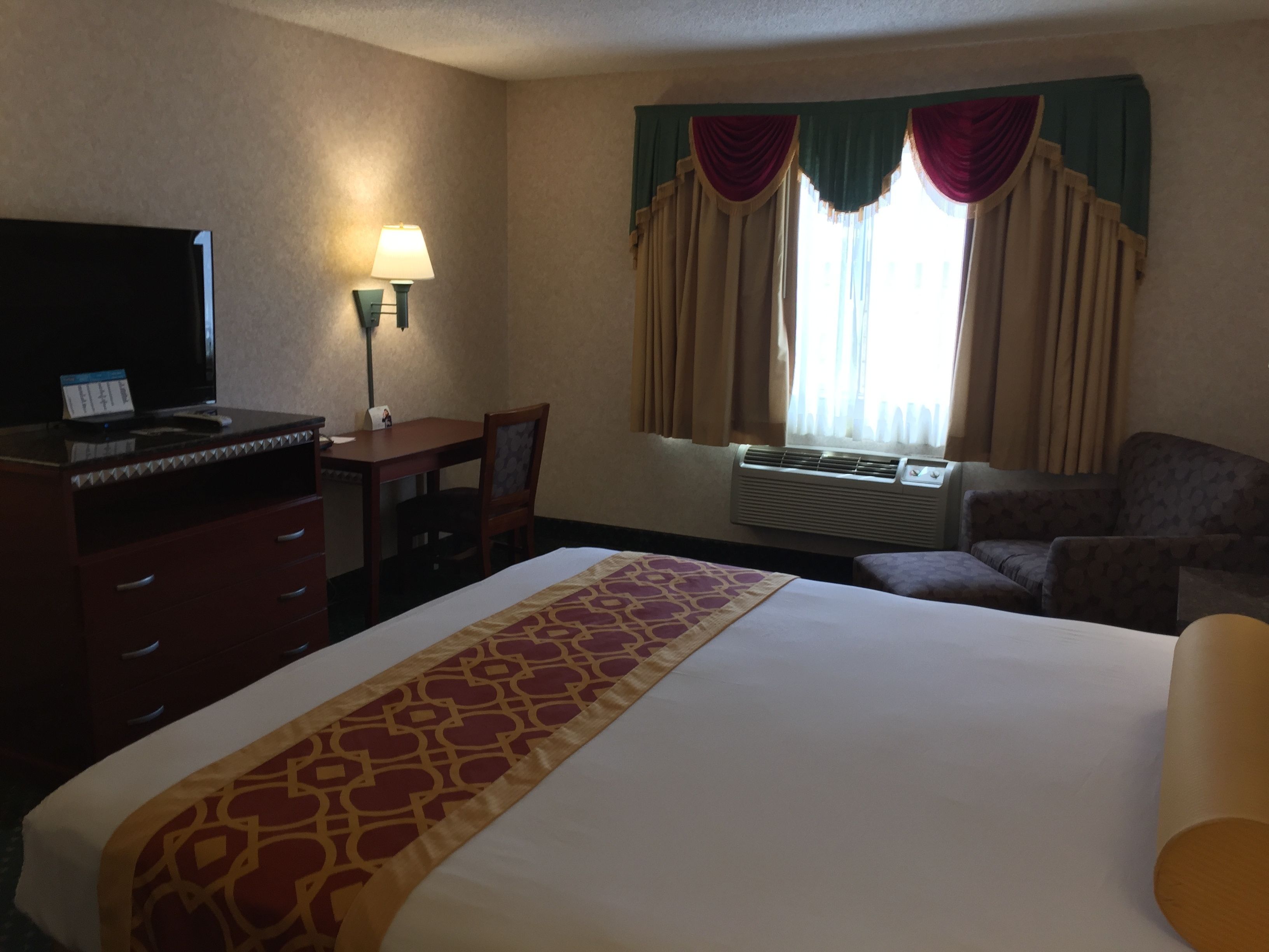 A hotel room with a bed, a desk, a chair, a television, and an air conditioning unit.