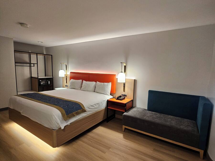 Habitación de hotel con cama, mesita de noche, lámpara, silla y un escritorio pequeño con teléfono.