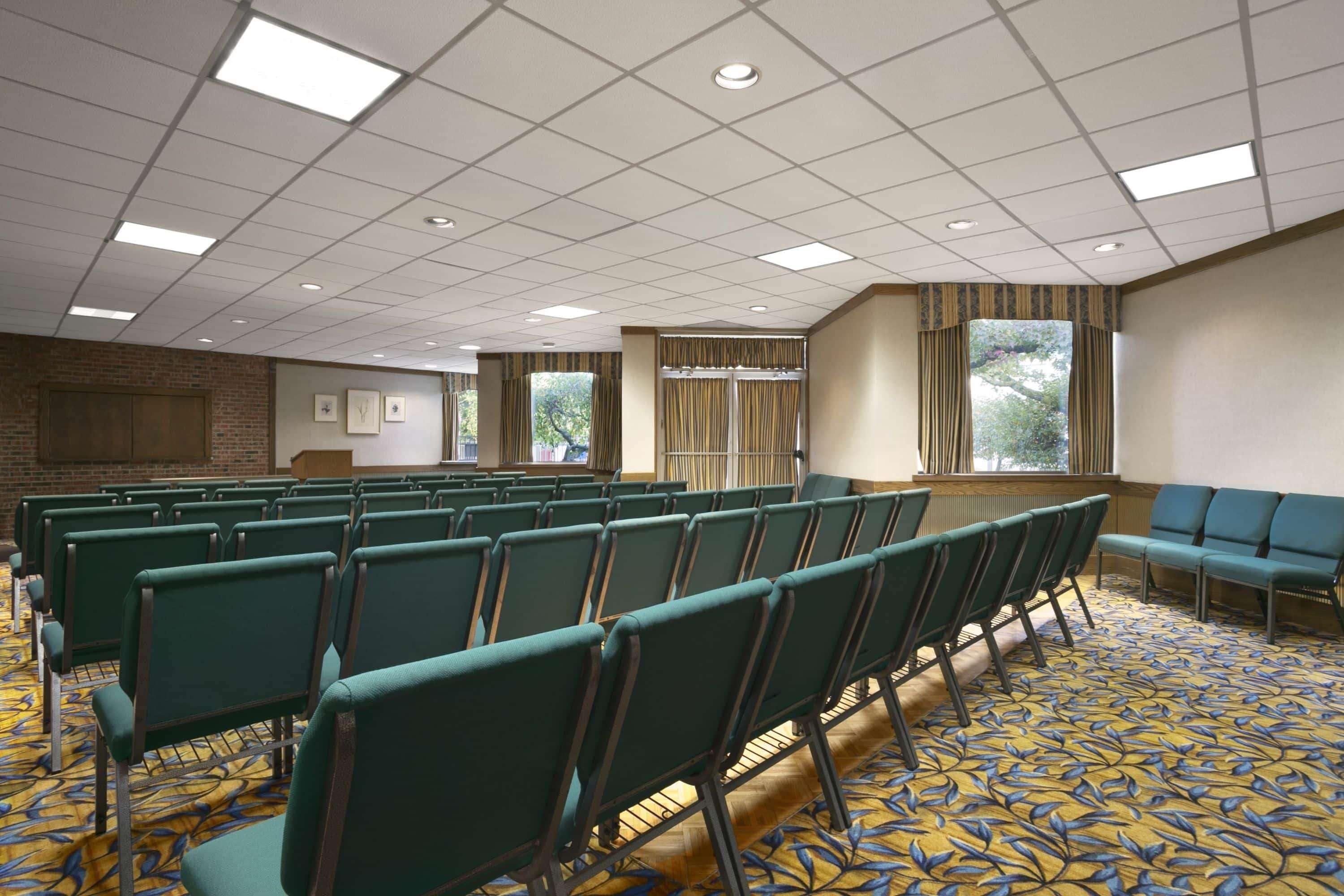 A conference room with rows of green chairs, a patterned carpet, and large windows with curtains.