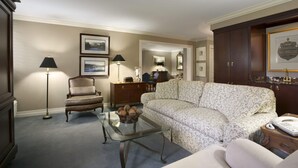 Un salon avec un canapé, un fauteuil et une table basse en verre. Des tableaux encadrés ornent le mur et une lampe est posée sur le sol.