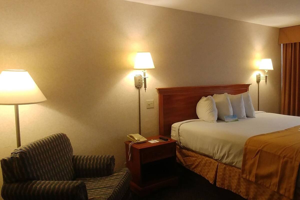 A hotel room with a bed, two bedside lamps, a chair, a nightstand with a phone, and a window with curtains.