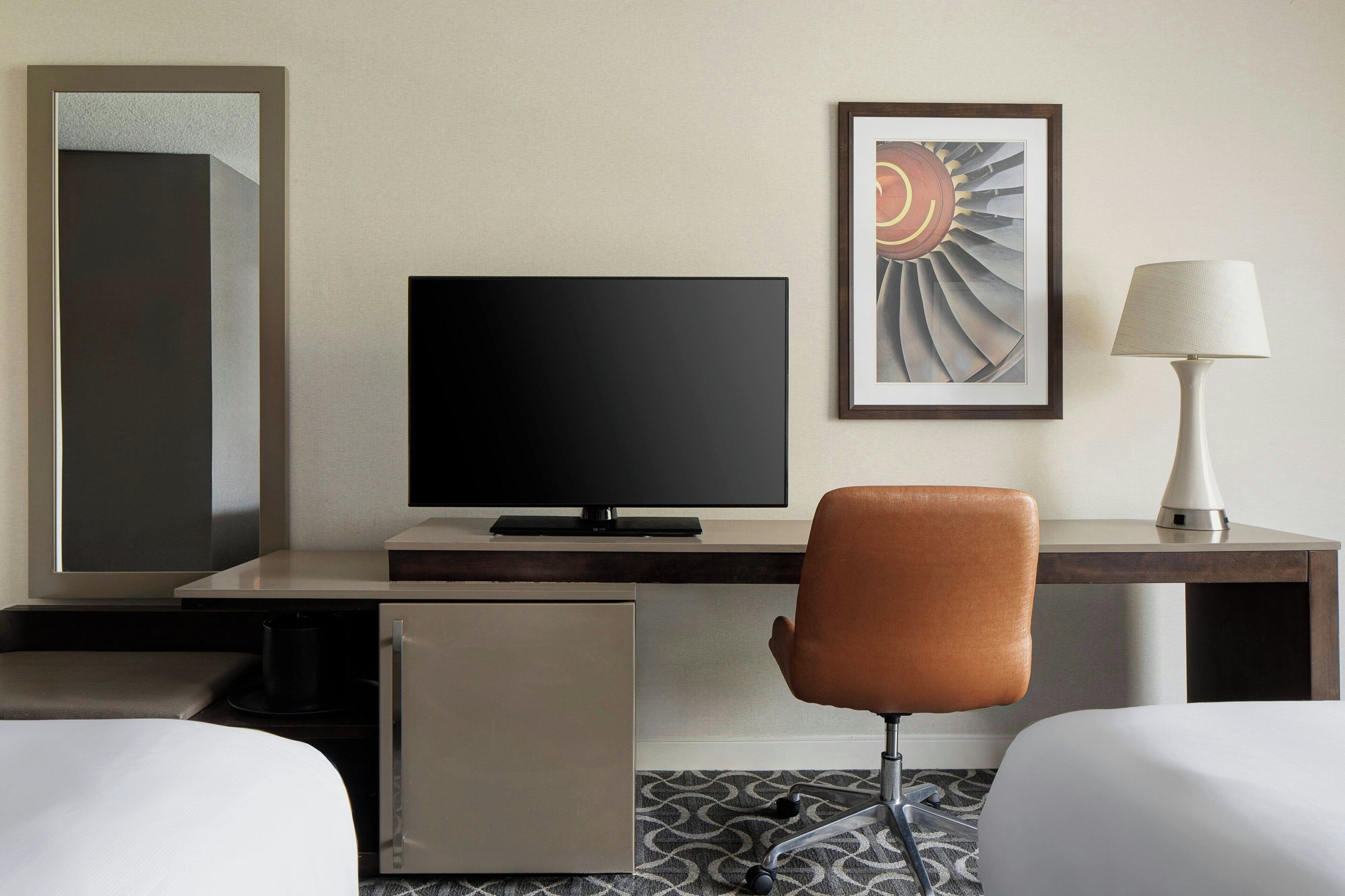 A hotel room with a bed, a desk with a computer, a chair, a lamp, and a framed picture on the wall.
