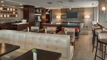 A modern hotel lobby with a bar area, seating arrangements, and a visible kitchen in the background.