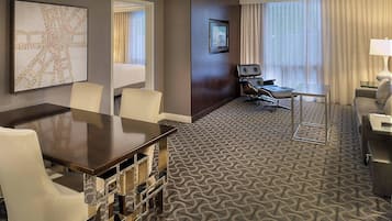 Habitación de hotel con un mapa grande en la pared, zona de comedor con mesa y sillas, espacio de estar con sofá y sillones, y un ventanal con cortinas.