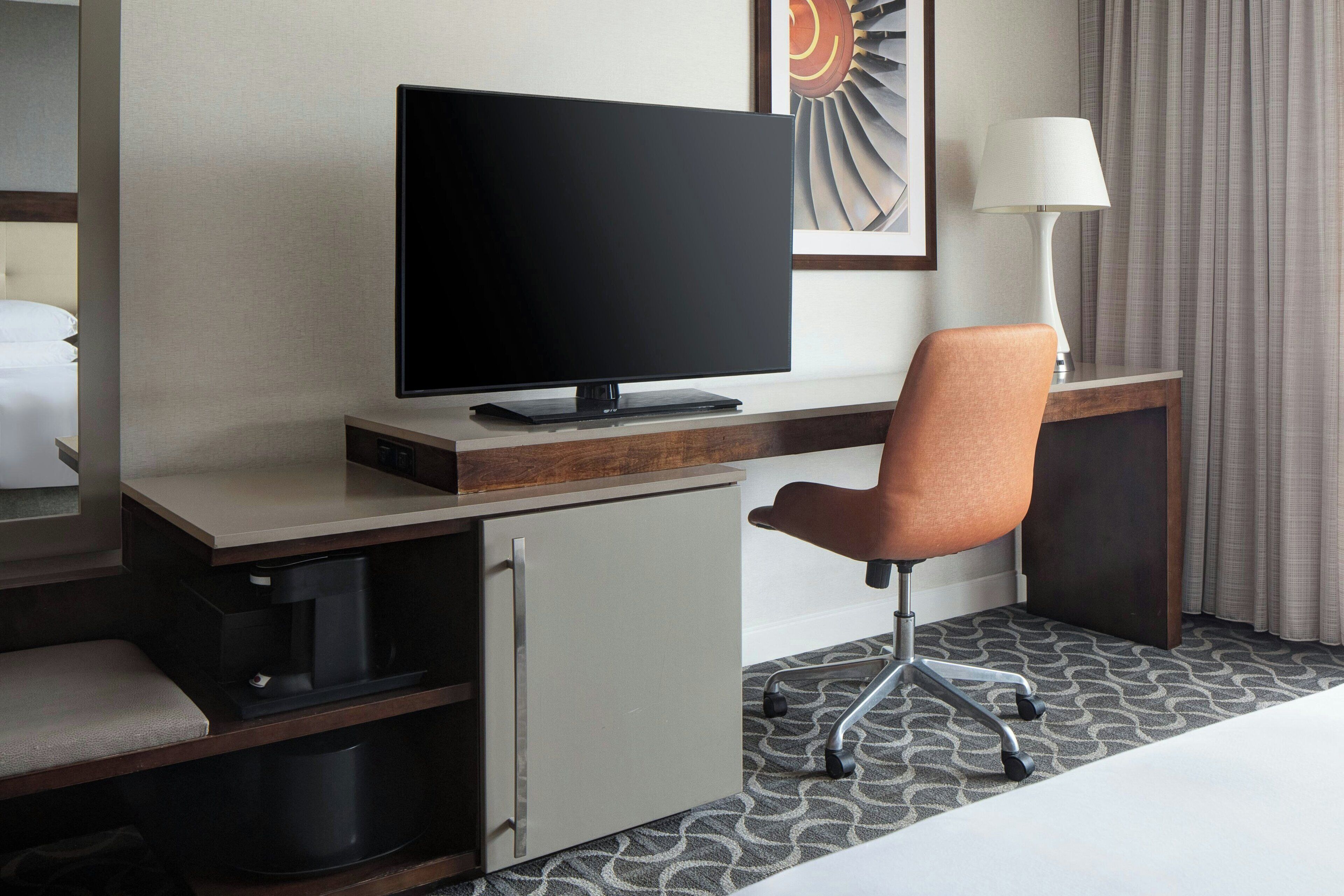 A hotel room with a flat-screen TV on a wooden desk, a brown office chair, a small cabinet, and a bed with white bedding.