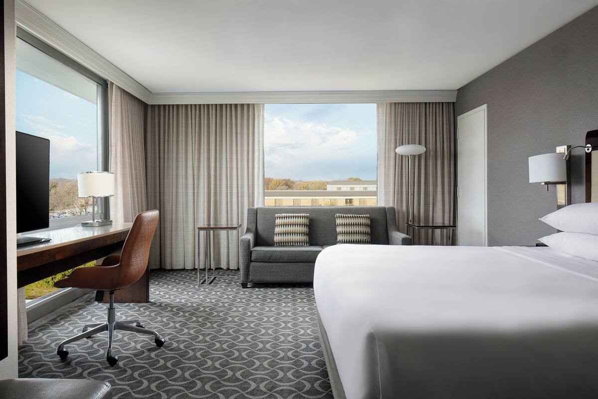 Habitación de hotel con una cama grande, un escritorio con silla, un sofá con almohadas, un televisor y una ventana con cortinas.