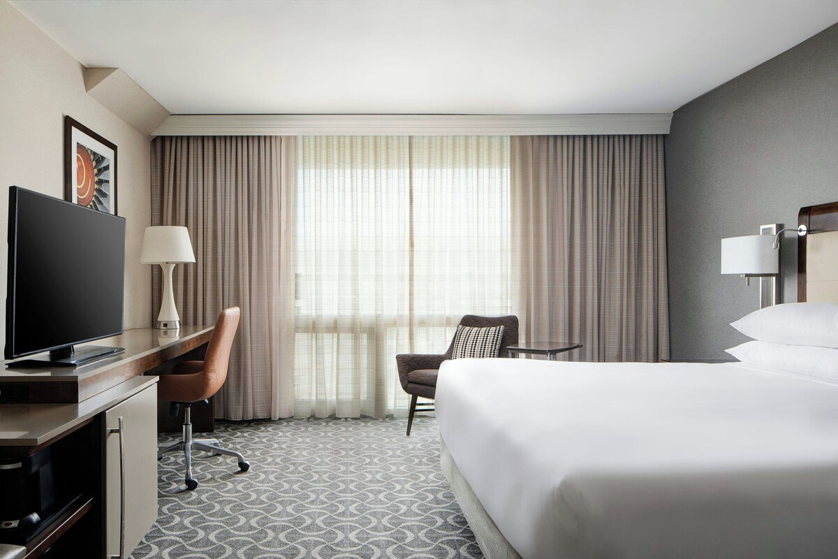 A hotel room with a bed, a desk with a TV, a chair, a lamp, and a window with curtains.