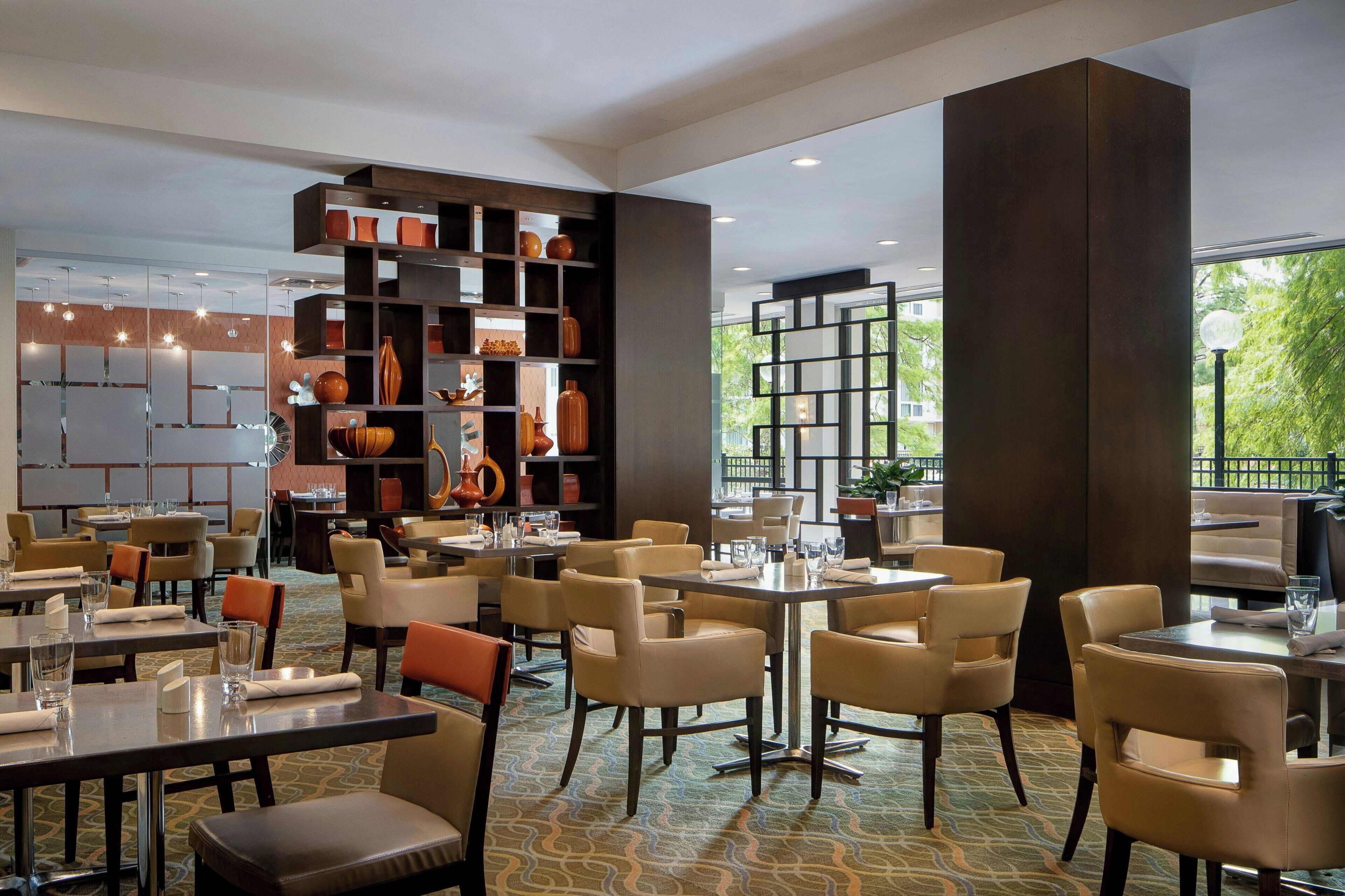 A spacious restaurant with multiple dining tables and chairs, a large bookshelf filled with decorative items, and large windows offering a view of the outdoors.