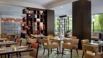 A spacious restaurant with multiple dining tables and chairs, a large bookshelf filled with decorative items, and large windows offering a view of the outdoors.