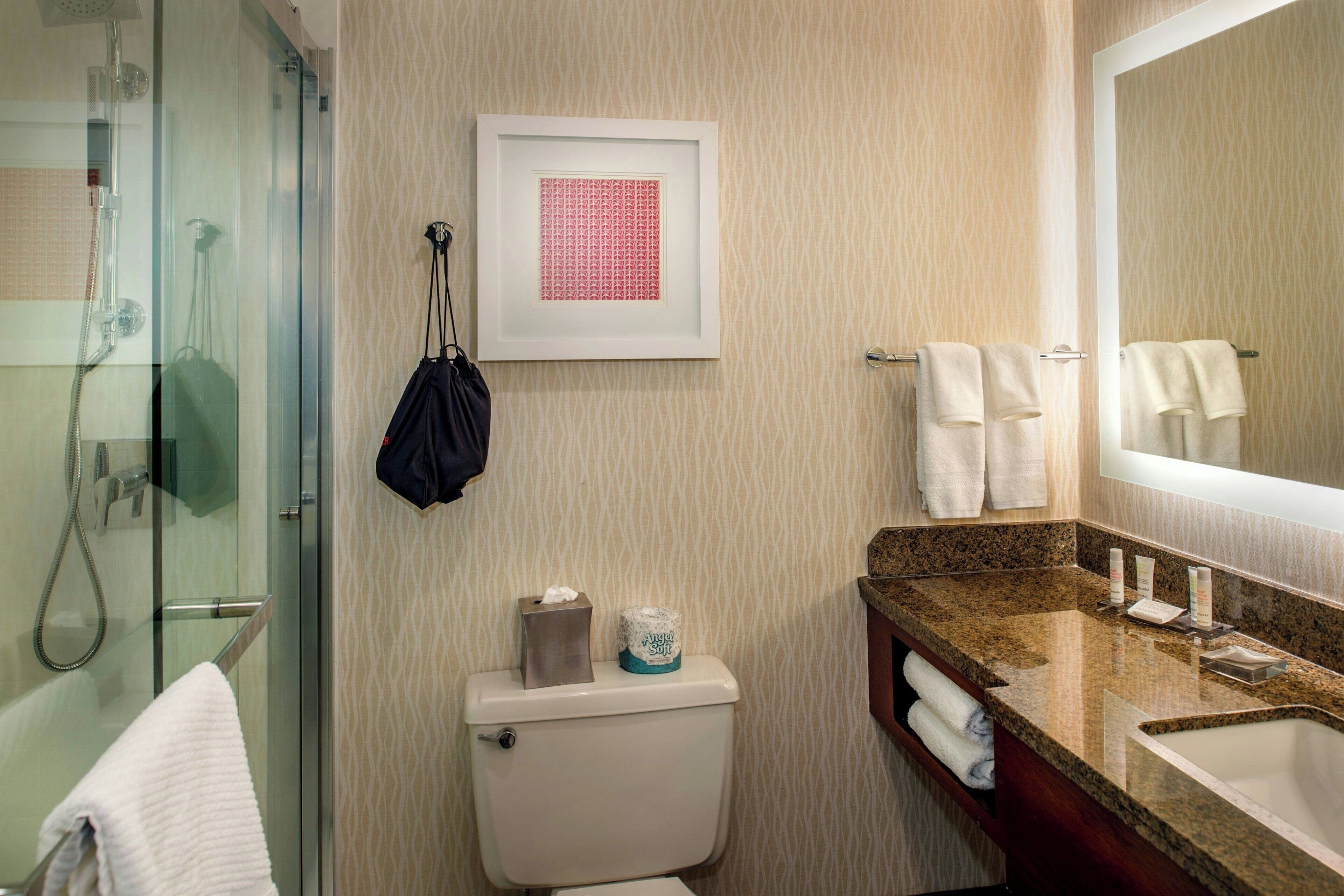 A bathroom with a glass shower enclosure, a toilet with a tissue box and toilet paper, a sink with a mirror, and towels on the wall.