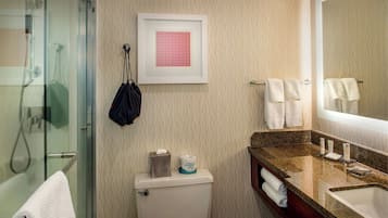 A bathroom with a glass shower enclosure, a toilet with a tissue box and toilet paper, a sink with a mirror, and towels on the wall.