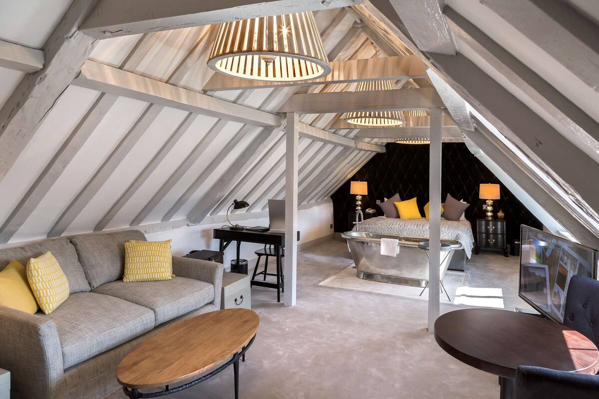 A cozy attic bedroom with a sloped ceiling, a sofa, a desk, a chair, a bed, and a bathtub.