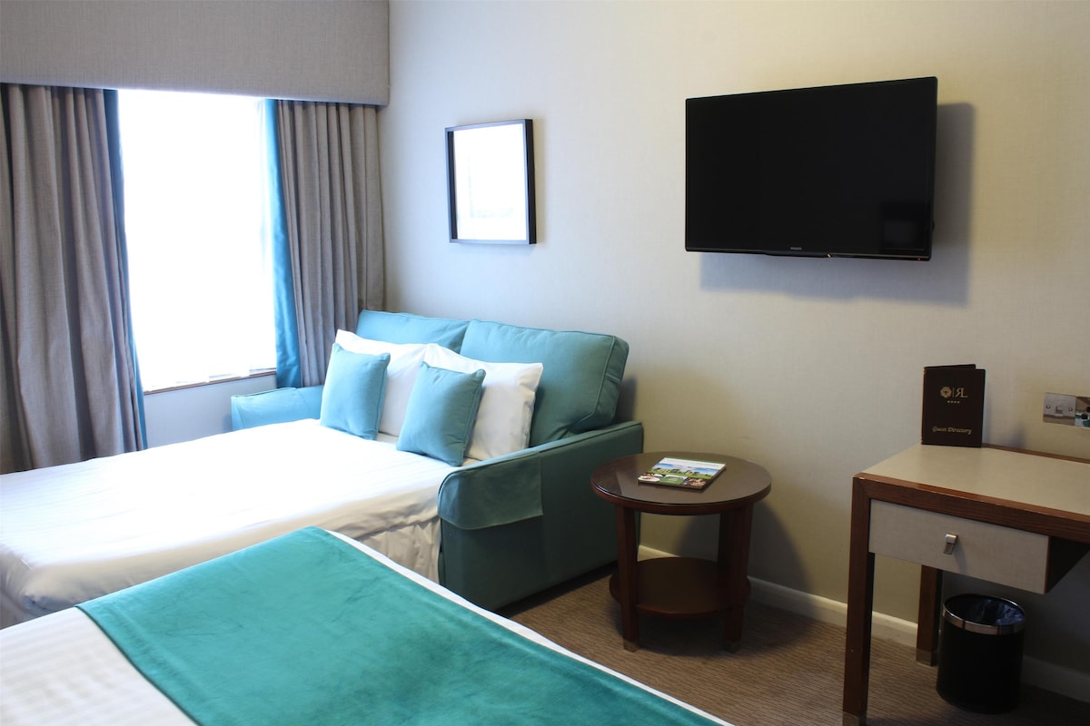 A hotel room with a bed, a sofa, a TV, a small table, and a trash bin.