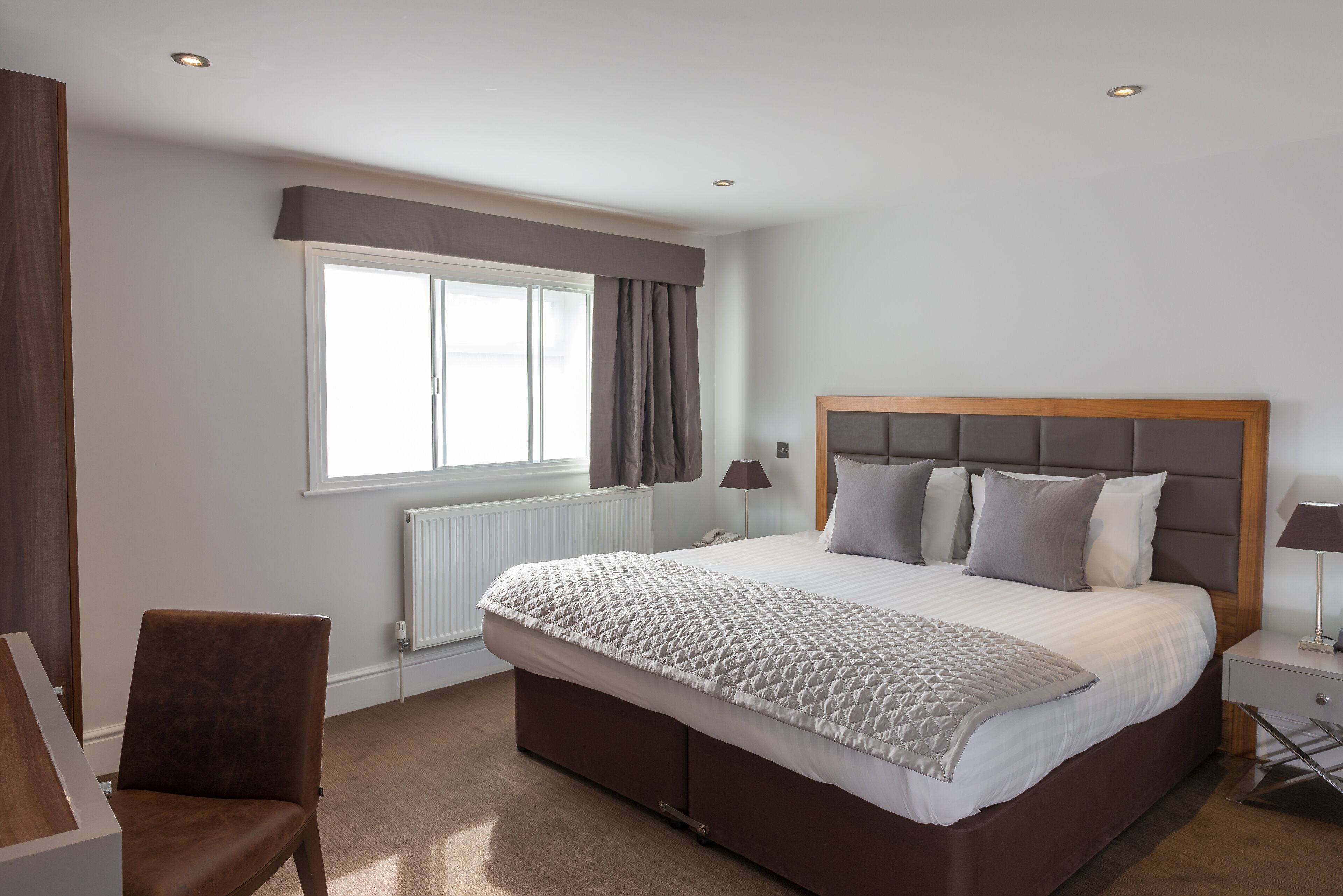 A modern bedroom with a large bed, a window with curtains, a chair, and a bedside table.
