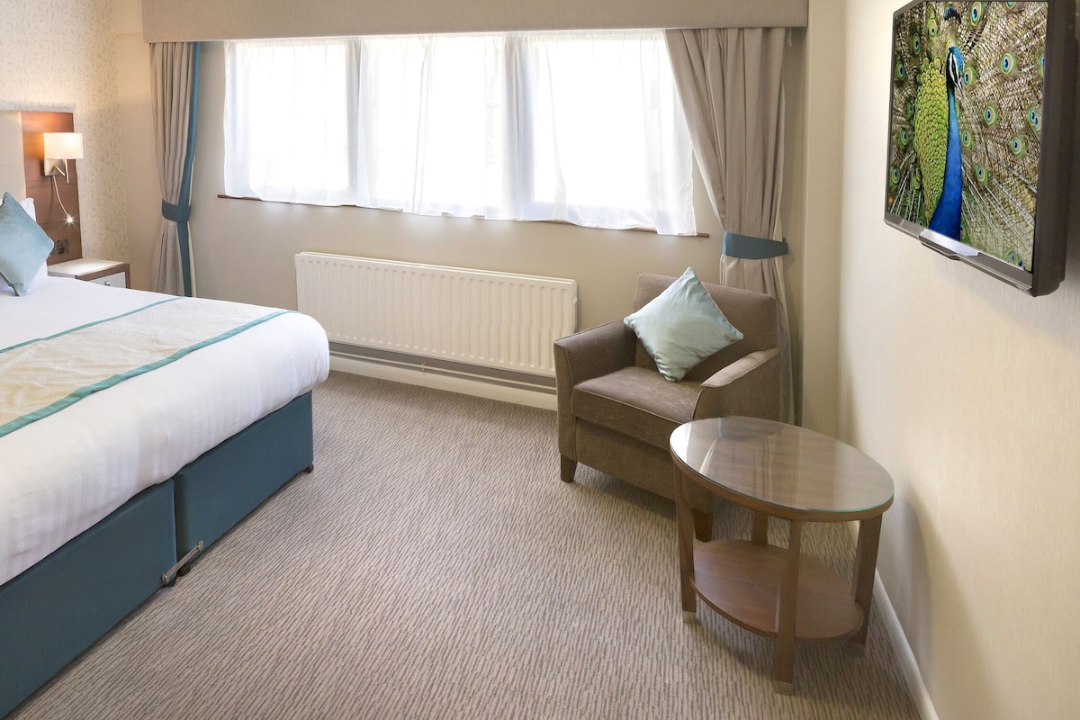 Habitación de hotel con una cama grande, mesitas de noche, una silla, una pequeña mesa, un televisor y una ventana con cortinas.