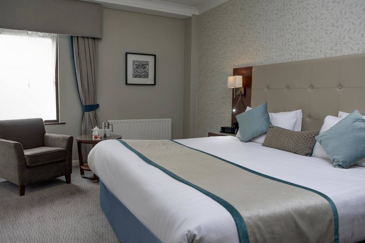 A hotel room with a large bed, a chair, a bedside table with a lamp, and a framed picture on the wall.