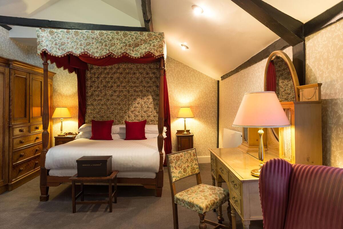 A four-poster bed with a canopy, two bedside lamps, a wooden dresser, a desk with a lamp, and a chair.