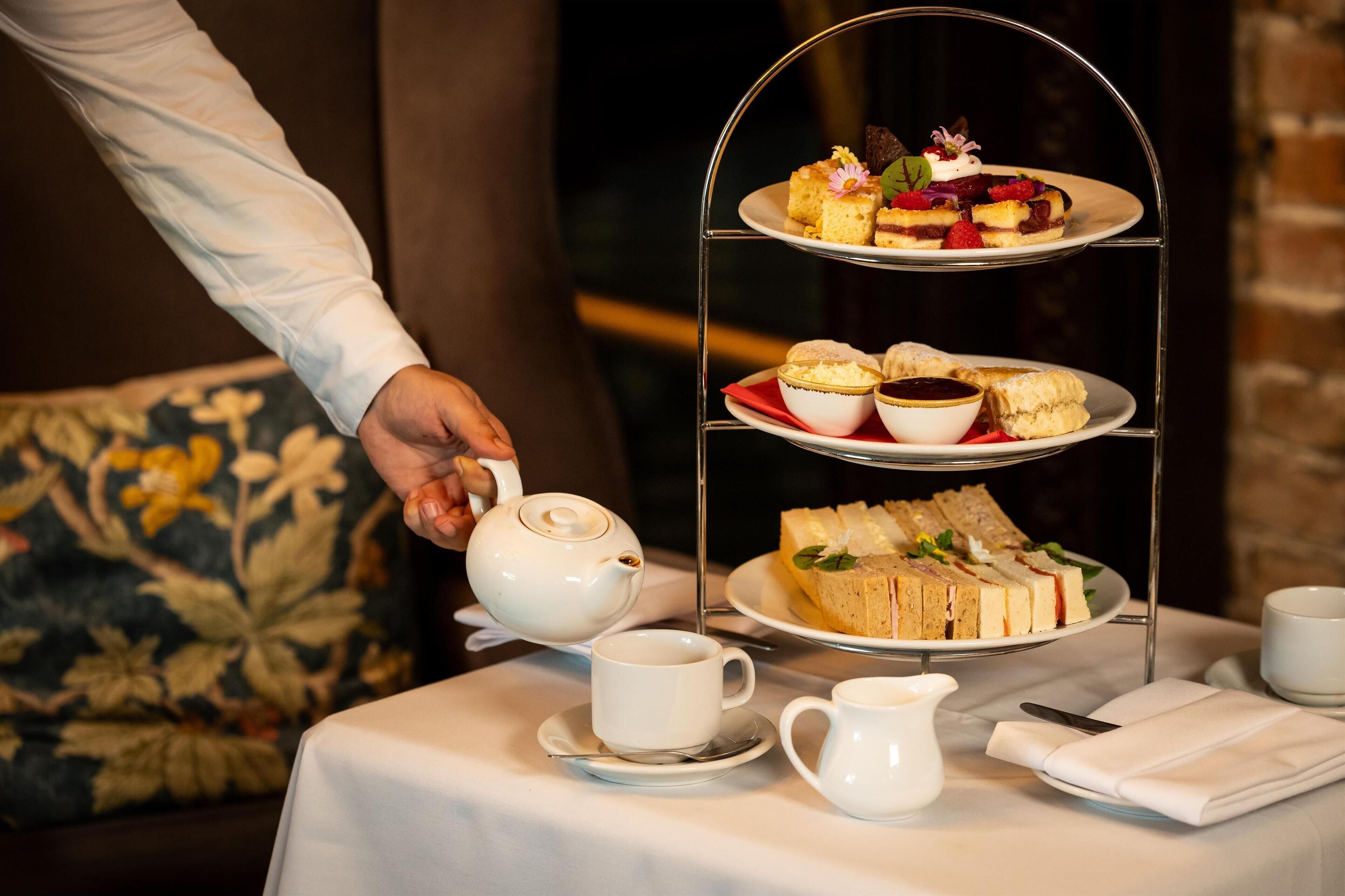 A tea service with a tiered tray of sandwiches and scones, a teapot, and cups on a table.