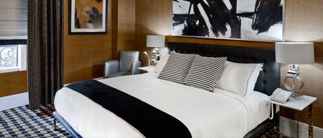 Una habitación de hotel con una cama grande, una mesita de noche con lámpara, un cuadro enmarcado en la pared y una ventana con cortinas.