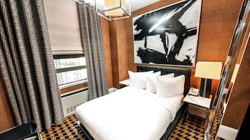 A hotel room with a bed, a bedside table with a lamp, a large abstract painting on the wall, patterned curtains, and a window with blinds.