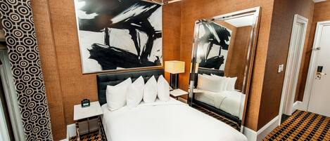 Una habitación de hotel con una cama grande, una mesita de noche con lámpara, un espejo amplio y una alfombra estampada.