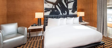 Habitación de hotel con una cama grande, una silla, una mesita de noche con lámpara, una obra de arte en la pared y una alfombra estampada.