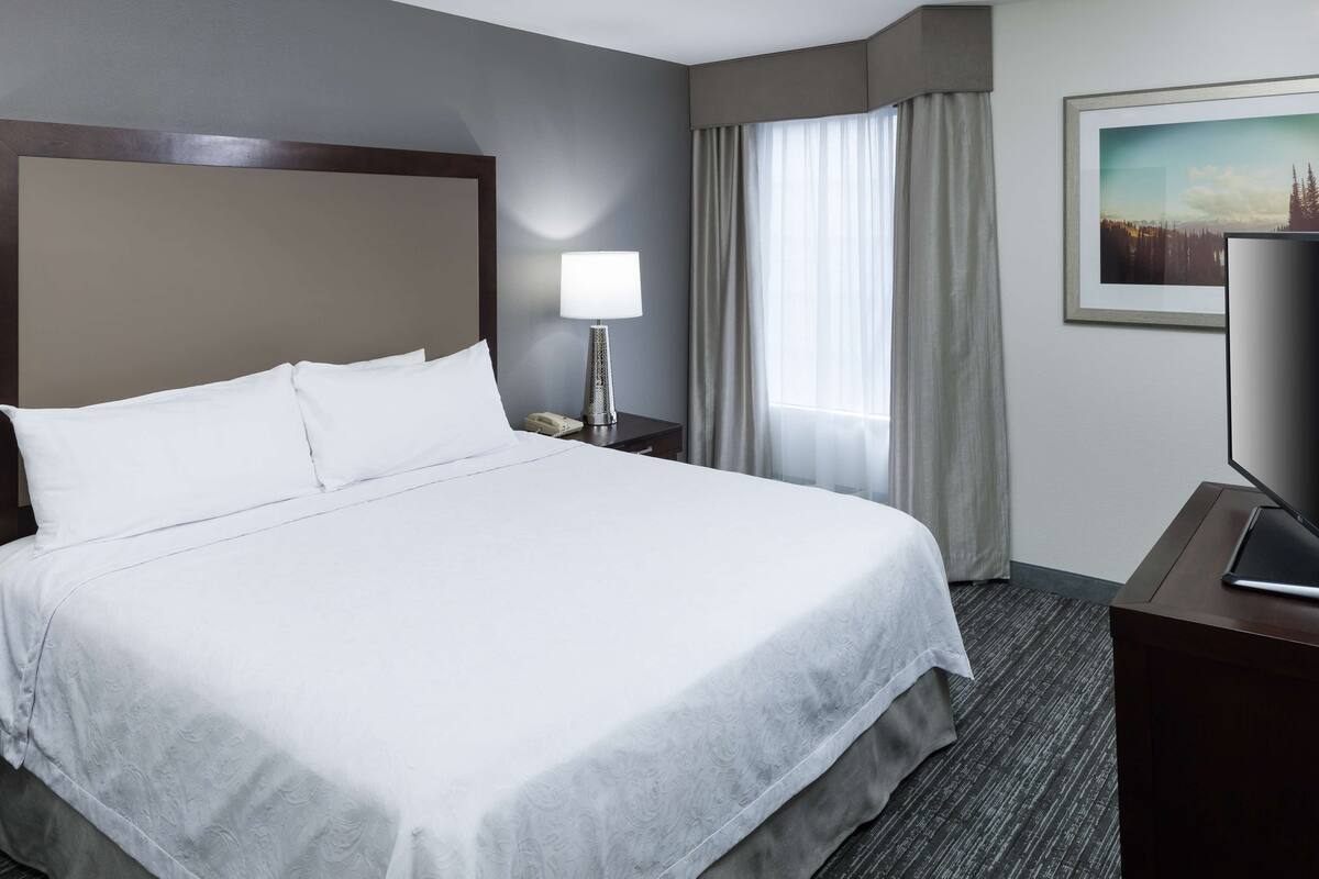 A hotel room with a large bed, a bedside lamp, a television, and a window with curtains.