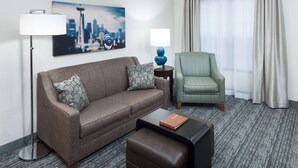 A hotel room with a brown sofa, a green armchair, a side table with a lamp, a coffee table, and a cityscape painting on the wall. - Homewood Suites by Hilton Seattle-Tacoma Airport/Tukwila (Tukwila)