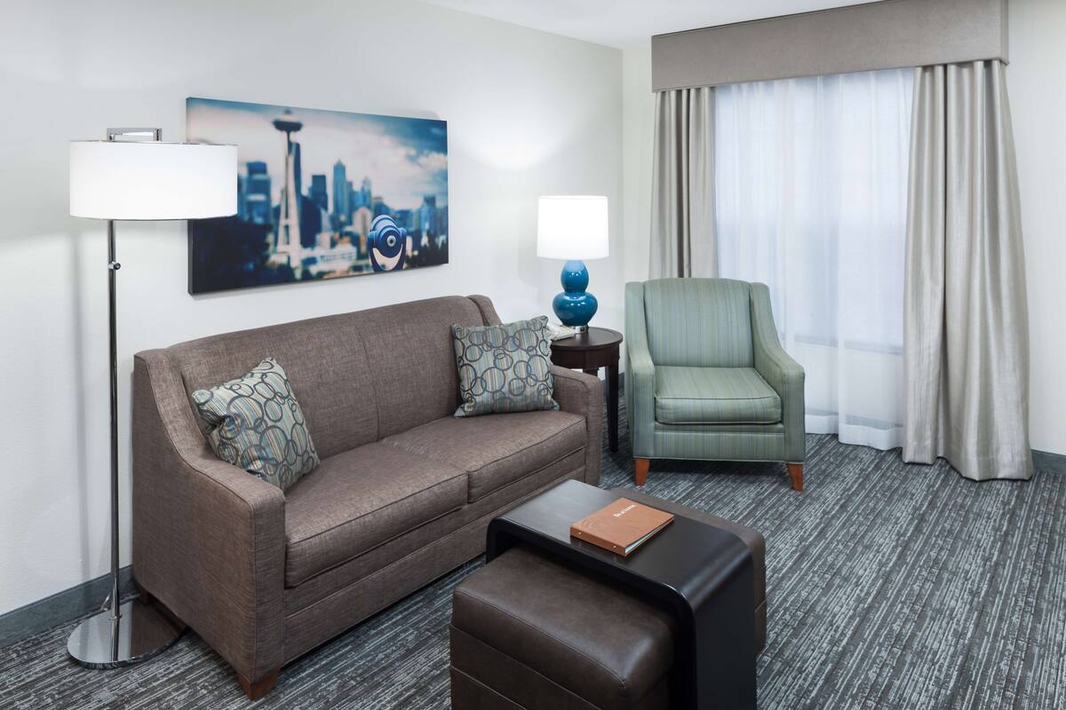 A hotel room with a brown sofa, a green armchair, a side table with a lamp, a coffee table, and a cityscape painting on the wall.