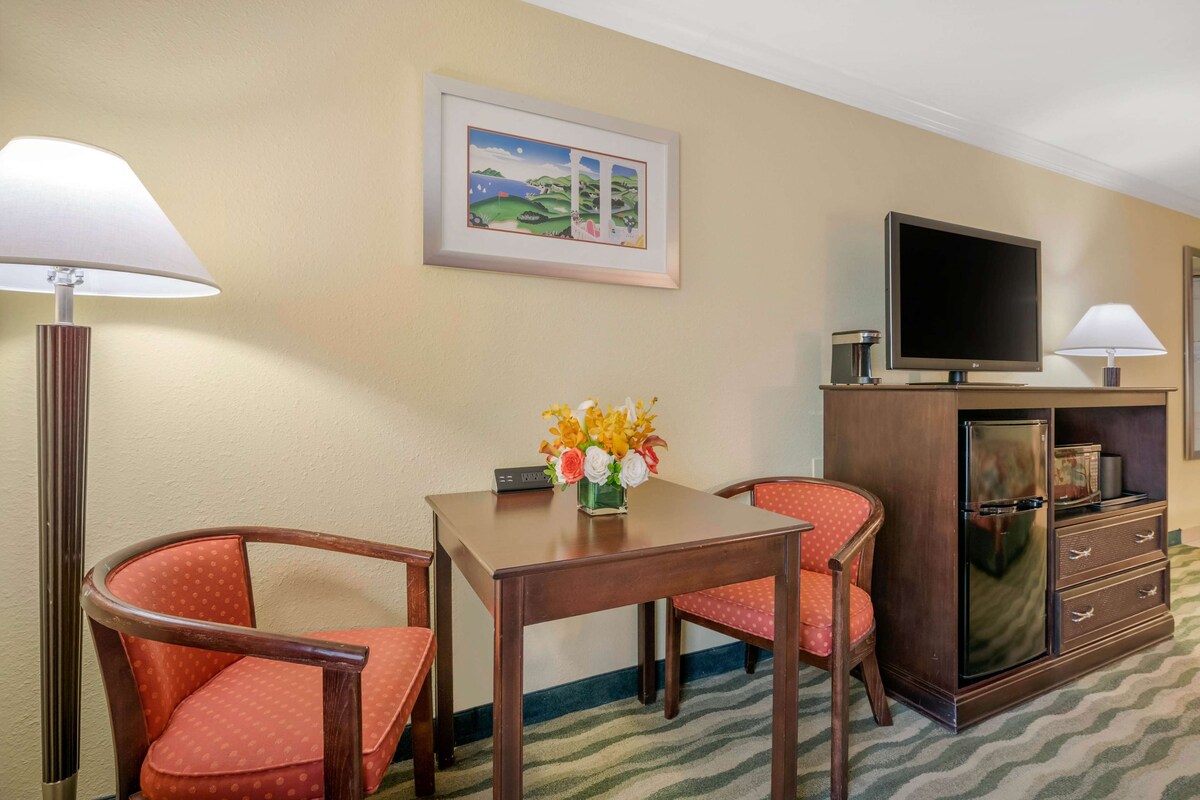 A hotel room with a wooden table, two red upholstered chairs, a television on a cabinet, and a framed picture on the wall.