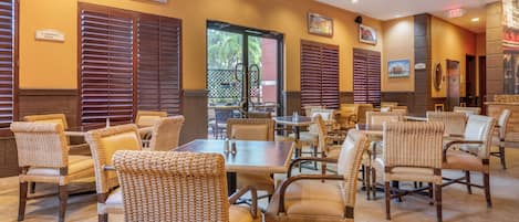 A restaurant with wicker chairs and tables, wooden blinds, and framed pictures on the walls.