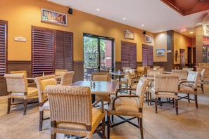 Een restaurant met rieten stoelen en tafels, houten jaloezieën en ingelijste foto’s aan de muren.