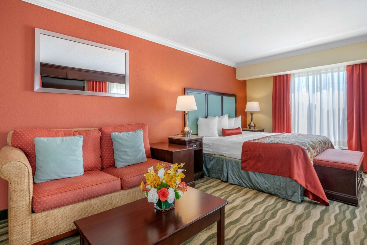 A hotel room with a sofa, a bed, a nightstand, a lamp, and a vase with flowers.