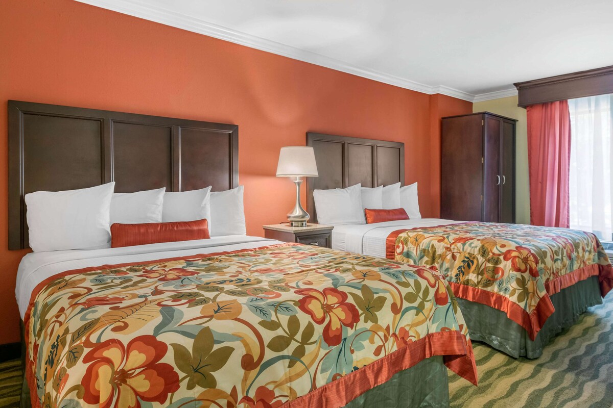 A hotel room with two beds, each with a floral bedspread, a nightstand with a lamp, and a wardrobe.