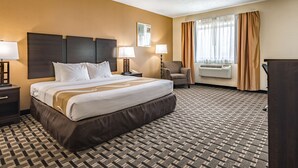 A hotel room with a large bed, two bedside tables with lamps, a chair, an air conditioning unit, and a window with curtains. - Quality Inn Schenectady - Albany (Schenectady)