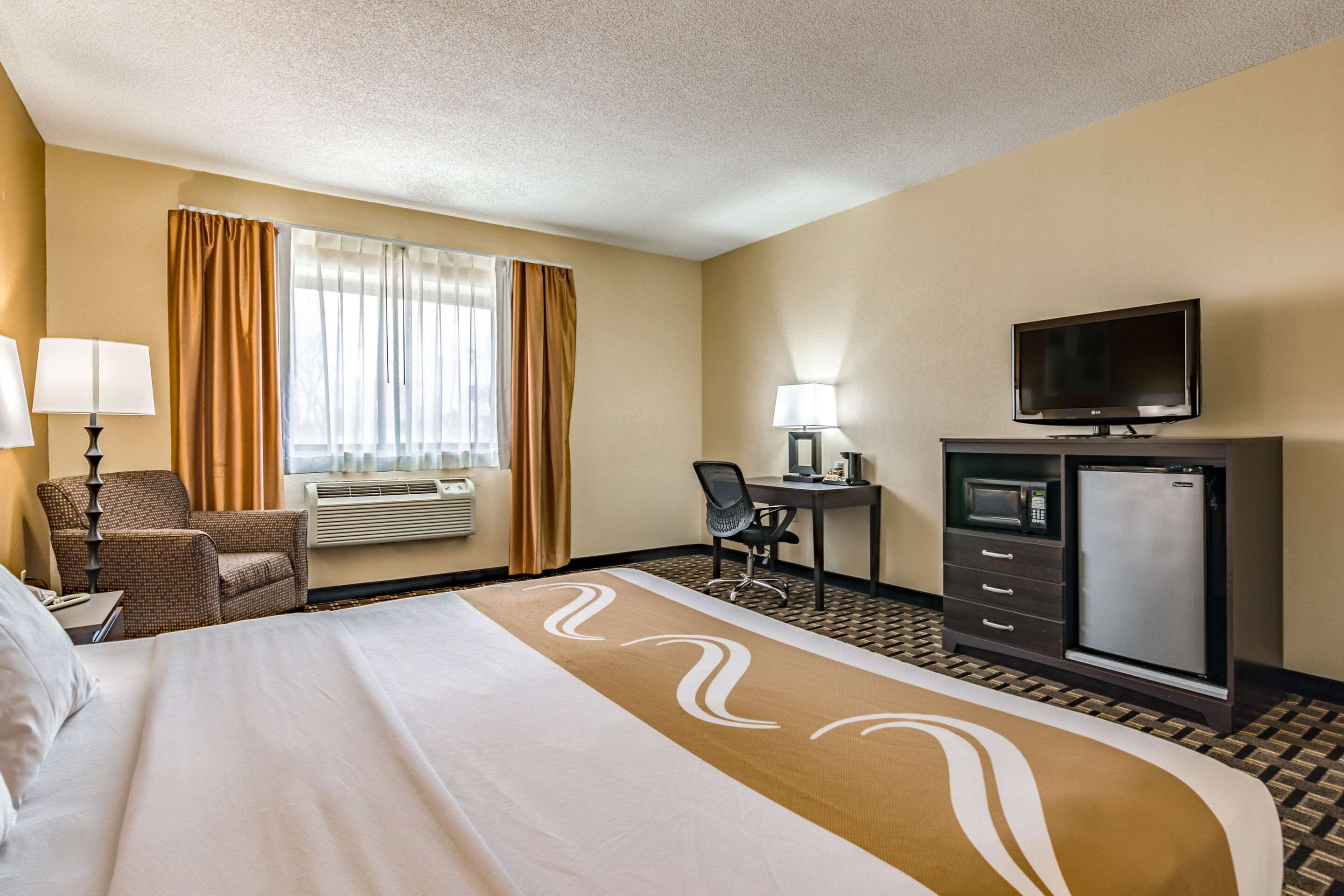 A hotel room with a large bed, a desk, a television, a refrigerator, and a chair.